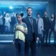 Diverse group of professionals standing in a blue-lit, futuristic corridor, some in lab coats and one in a suit, facing the camera.