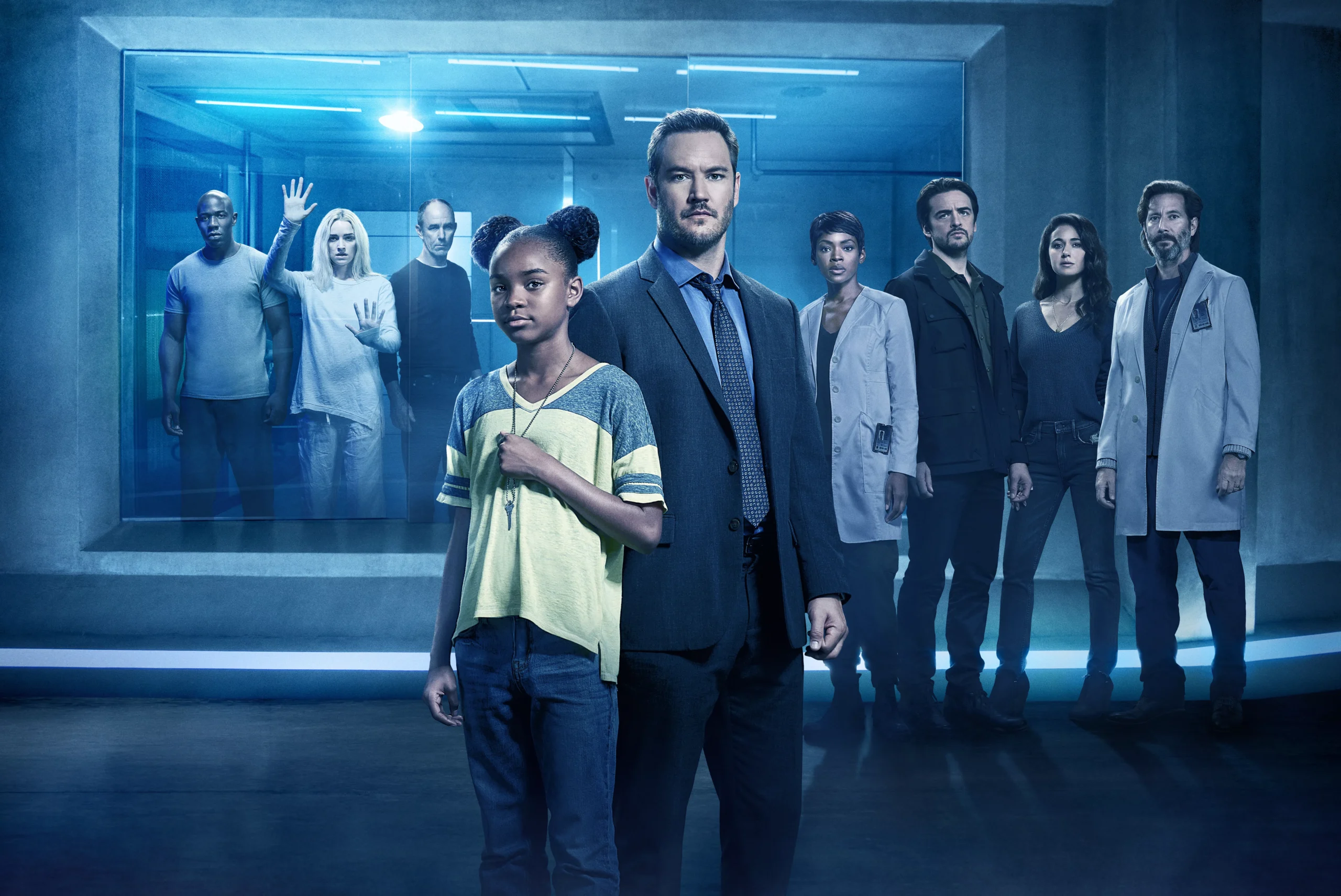 Diverse group of professionals standing in a blue-lit, futuristic corridor, some in lab coats and one in a suit, facing the camera.