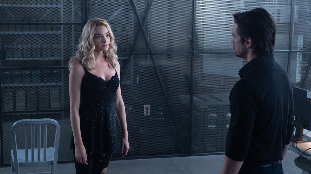 Two people stand in a dim industrial setting, facing each other as a woman in a black dress talks to a man in a dark shirt against a metal mesh backdrop.