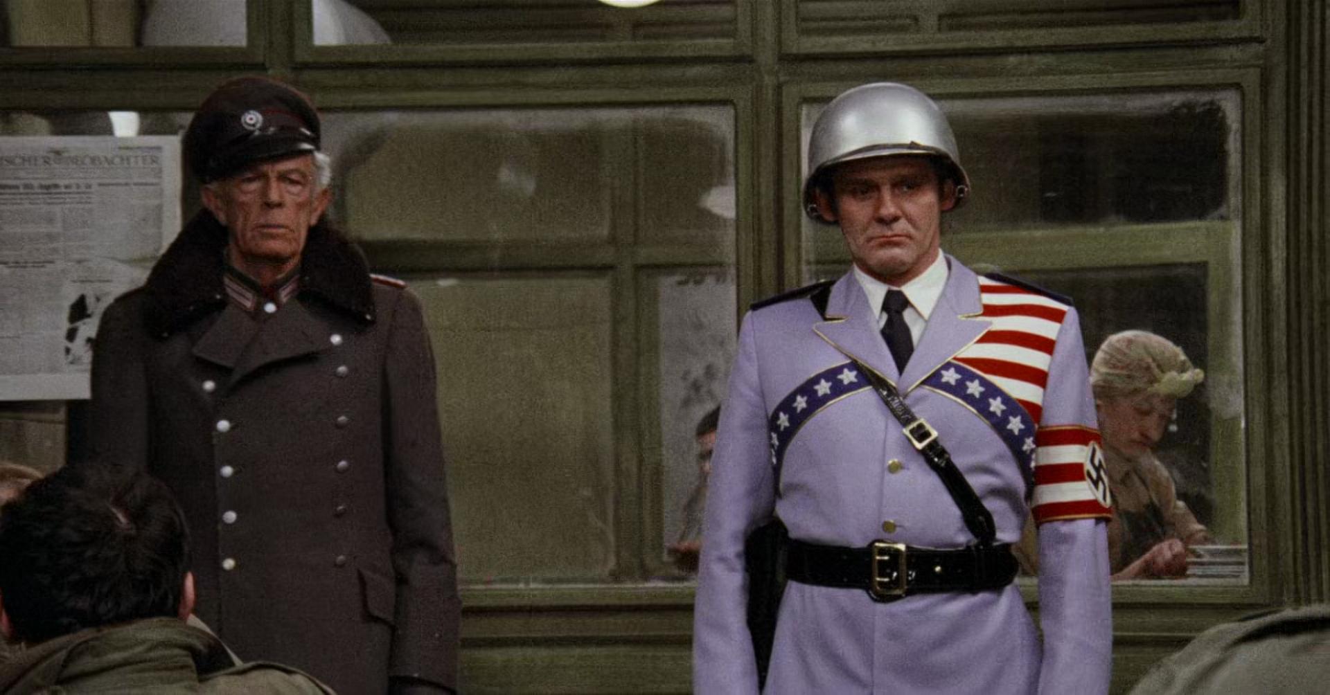 rzeznia-nr-5 | film.org.pl Two men in period military uniforms stand at a station or doorway; one wears a pale purple uniform with a helmet and a star-spangled sash, the other in a dark coat and peaked cap, both looking ahead.