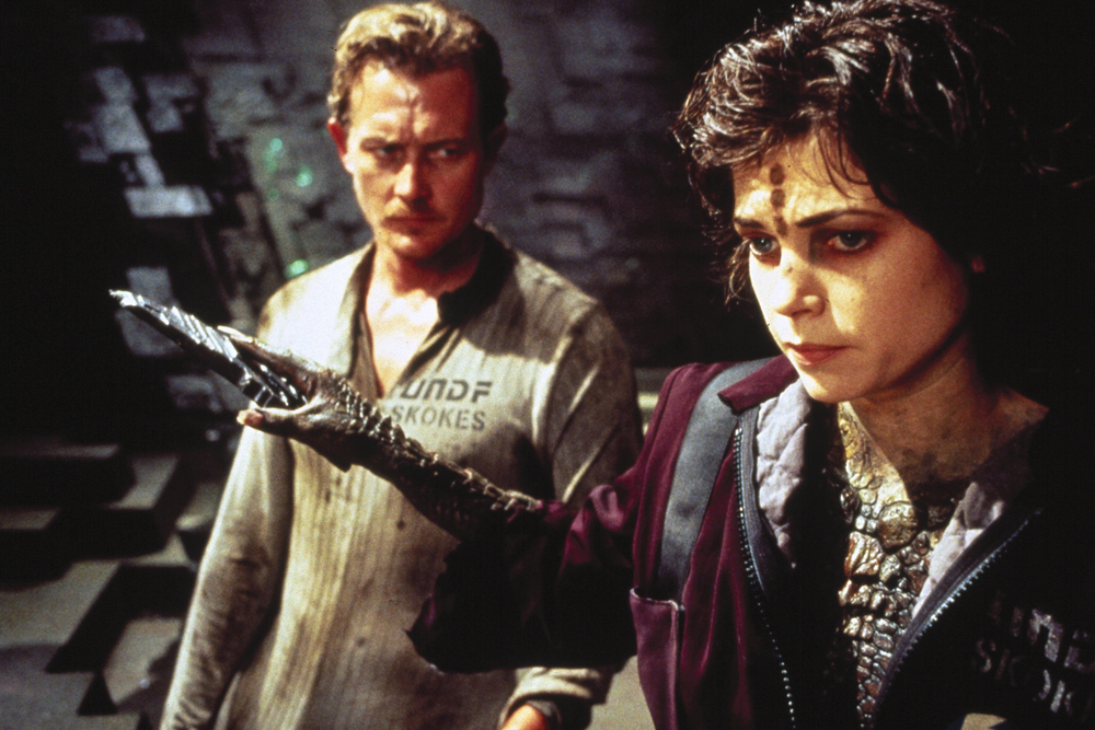 Woman with a metallic prosthetic arm extended forward, wearing a burgundy jacket and ornate necklace, as a concerned man in a beige shirt watches in a dark, industrial setting.