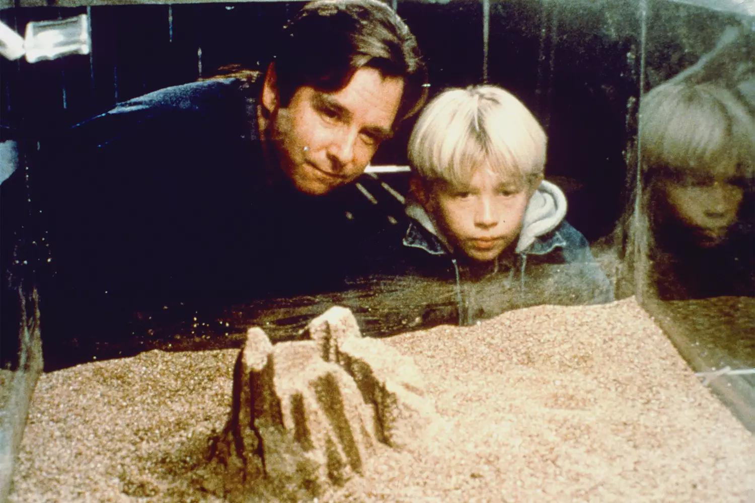 Man and young boy lean close together, looking at a pink sand sculpture inside a glass display case.