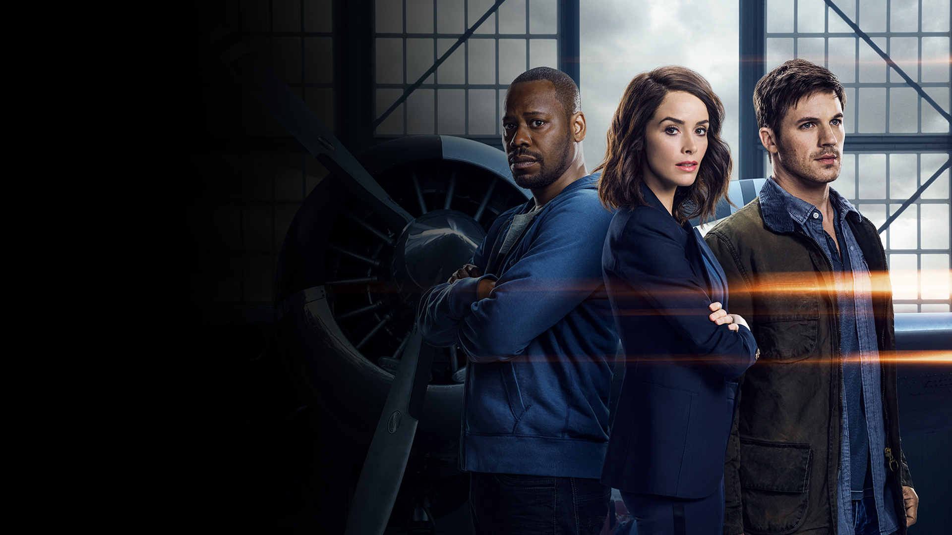 Three aviation professionals stand with arms crossed in a hangar, a jet engine and large windows behind them, suggesting a maintenance or engineering team.