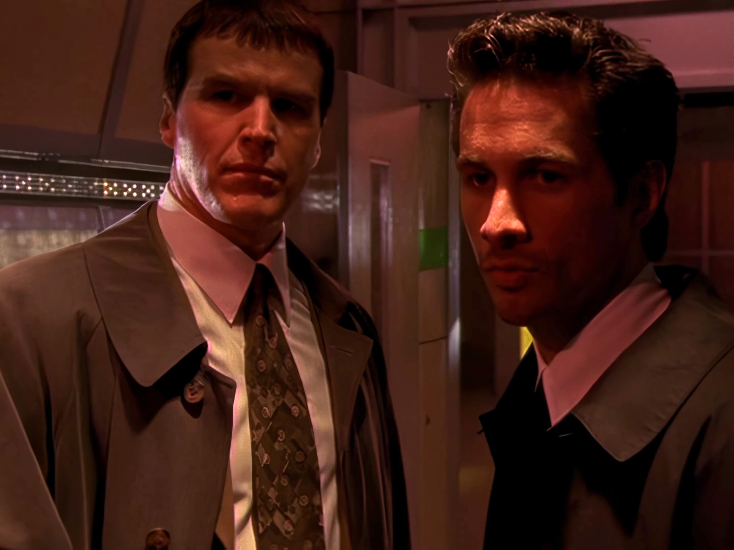 Two men in formal suits stand side by side in a dimly lit room, looking at the camera with serious expressions.