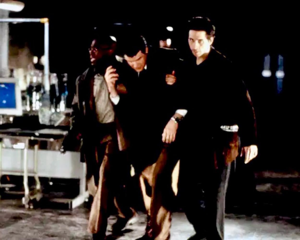 Three men in dark clothing walk together at night near industrial equipment.