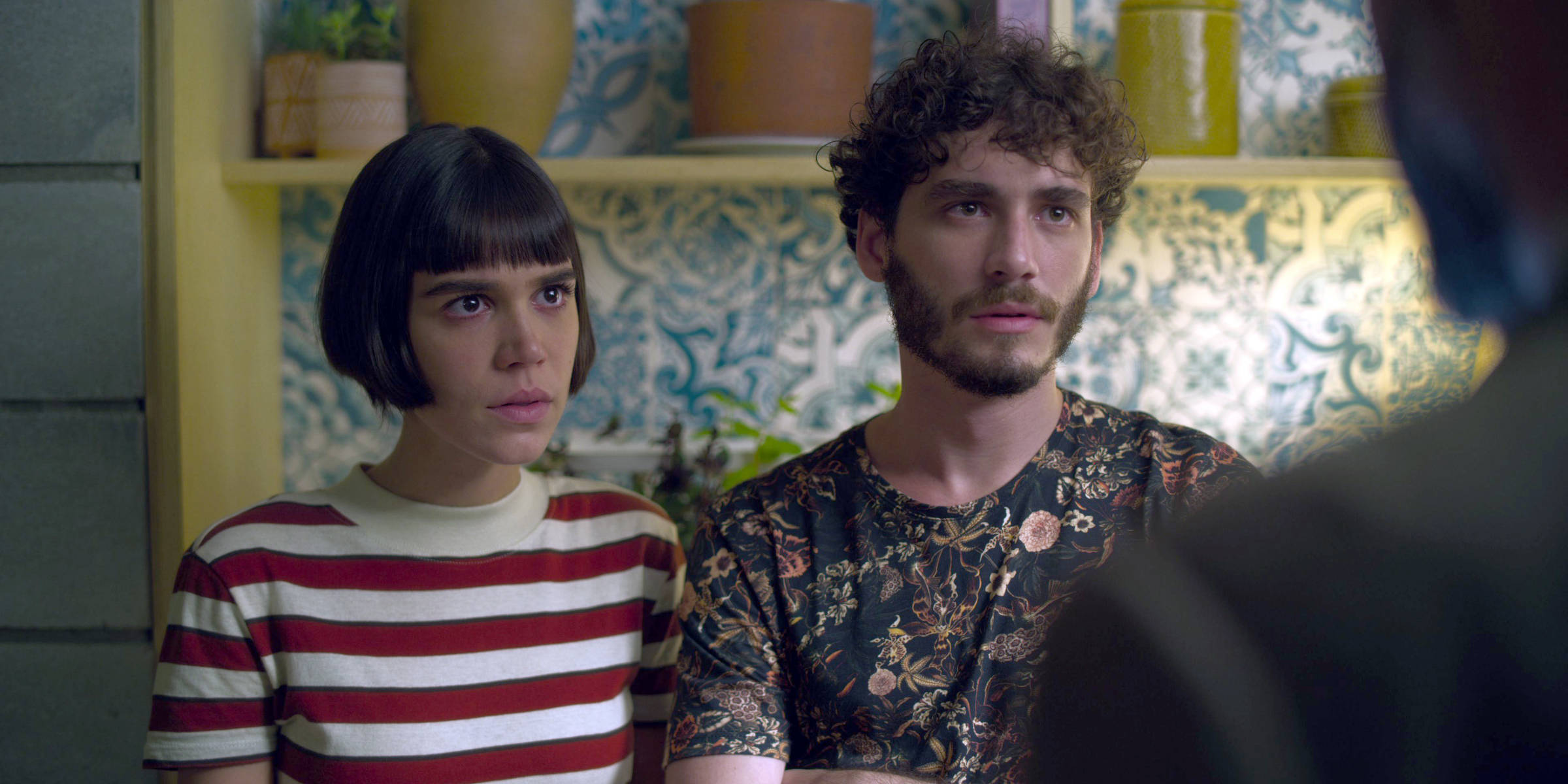 wszechwiedzacy | film.org.pl Two young adults sit side by side, listening intently to someone off-camera; the woman wears a red-and-white striped shirt and the man wears a floral-patterned shirt, in a colorful, patterned room.