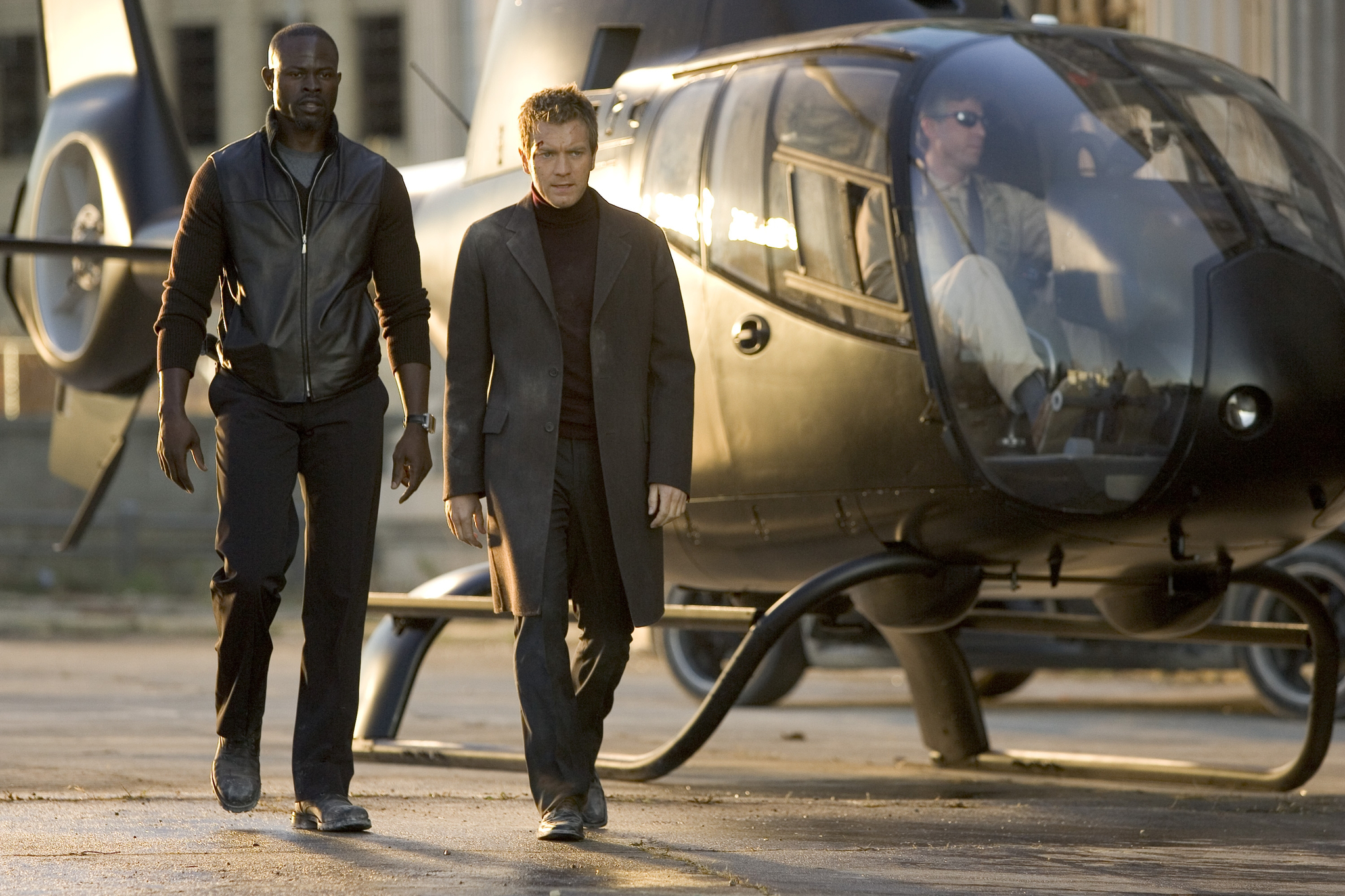 Two men walk toward the camera away from a gray helicopter on a tarmac at sunset, one in a black leather vest and the other in a long coat.