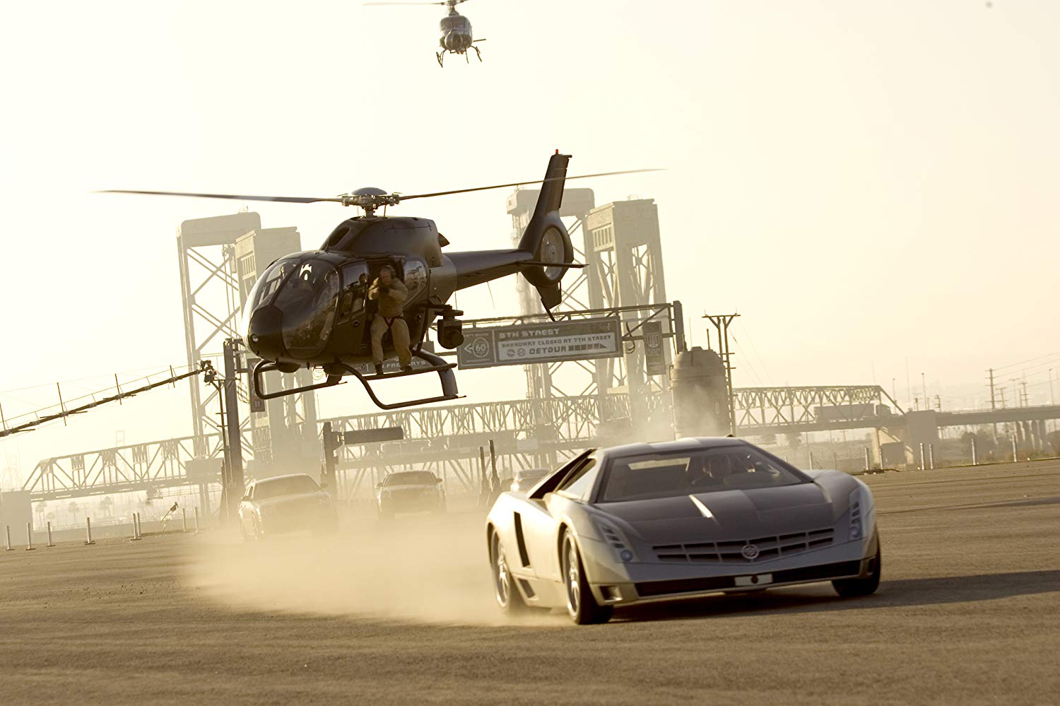 Low-flying helicopter hovers beside a silver sports car on a dusty runway with industrial bridges in the background.