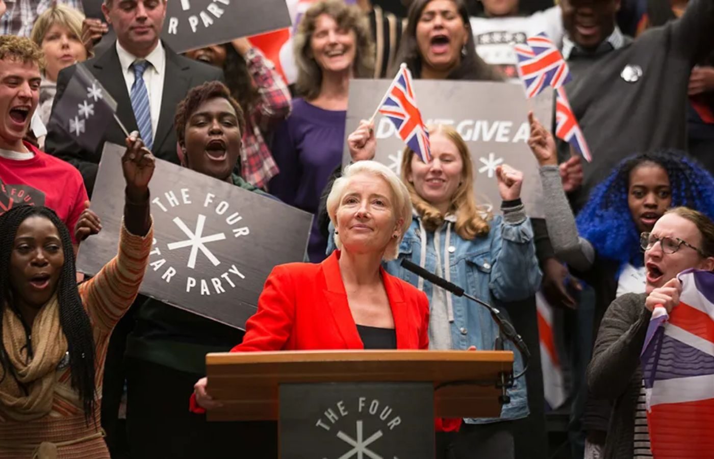 Politician in a red blazer speaks at a podium while a cheering crowd waves flags and holds signs that read 'The Four Star Party'.
