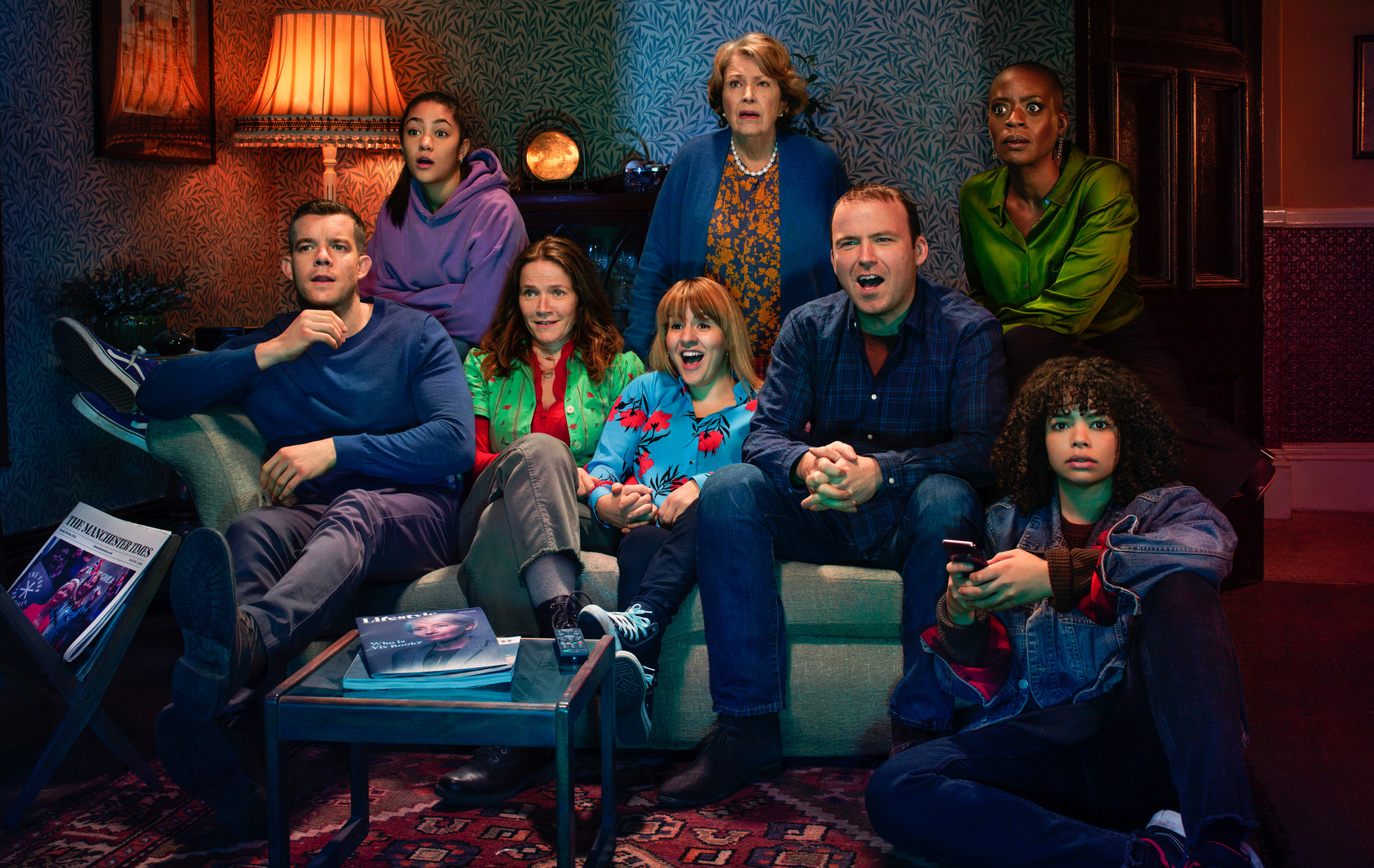 Group of seven people in a cozy retro living room, seated on a sofa and ottoman, reacting with surprise to something off-camera.