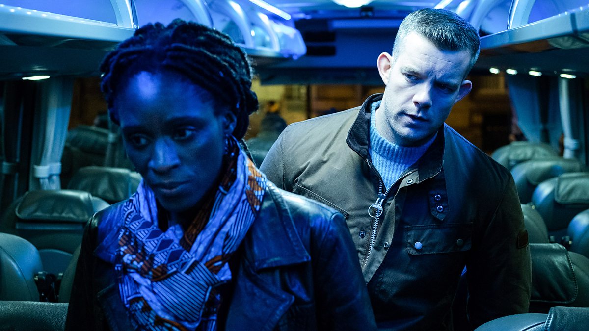 Two adults sit on a dimly lit bus with blue neon lighting, looking wary or lost in thought as they face forward.
