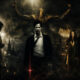 Dramatic urban fantasy scene: four people stand in a dark, smoky cityscape beneath a giant winged figure, with a central man in a white shirt and black coat and candles in the foreground.
