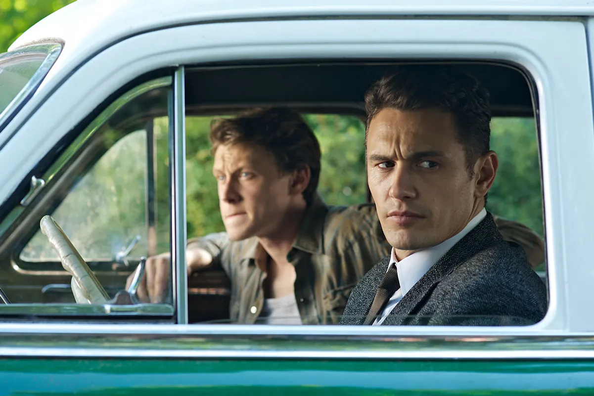 Two men in a vintage car: a suited passenger with a serious expression and a casually dressed driver.