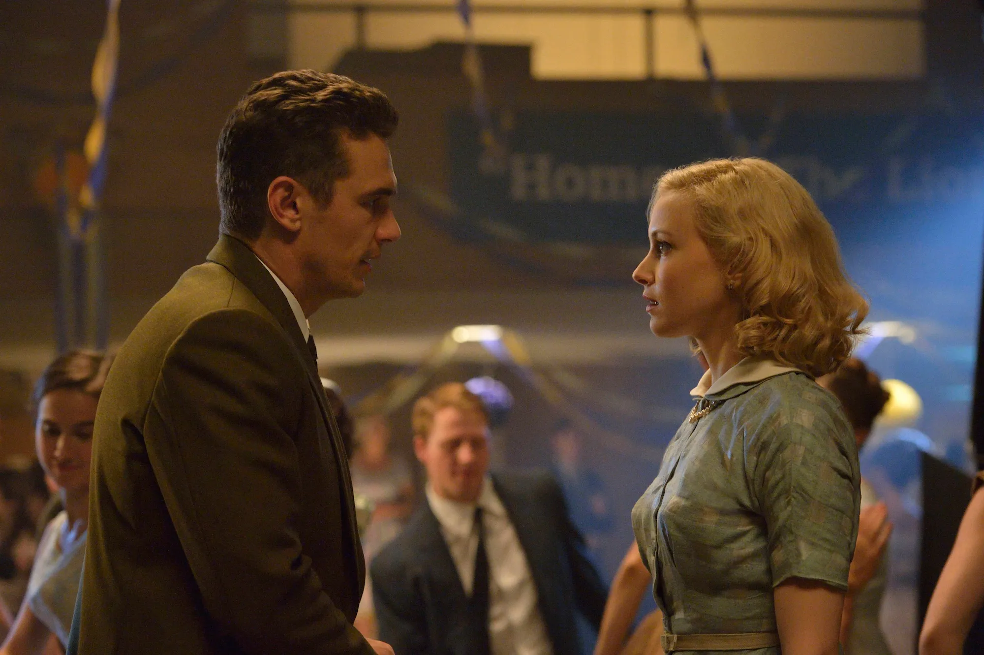 Man in a brown suit and a blonde woman facing each other in a dimly lit social event, with people in the background.