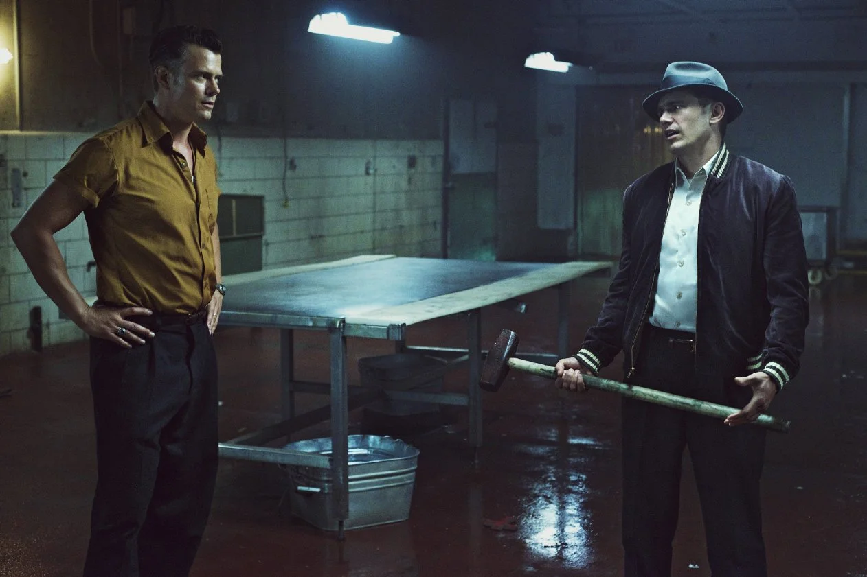 Two men in a dim warehouse-like room; one in a mustard shirt with hands on hips, the other in a hat and jacket holding a large hammer.