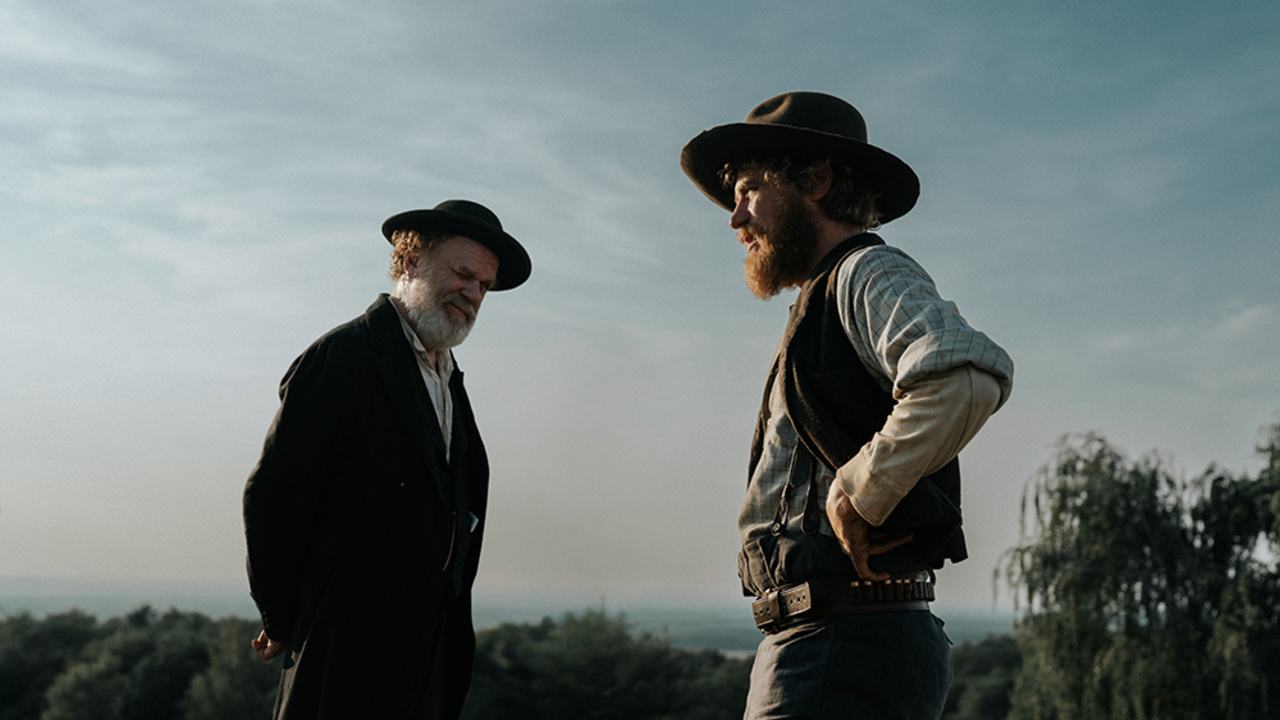 Two bearded men in vintage Western outfits and wide-brim hats talk outdoors in a rural landscape.