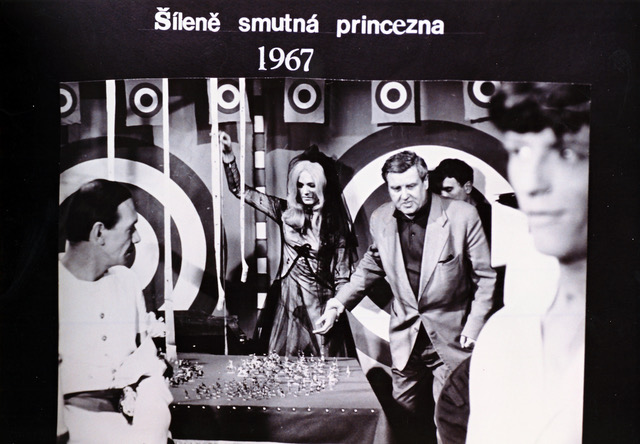 Black-and-white still from the 1967 film 'Šíleně smutná princezna' showing a man in a suit reaching toward a table with scattered small objects, with a woman in a dramatic outfit and other figures in the background.