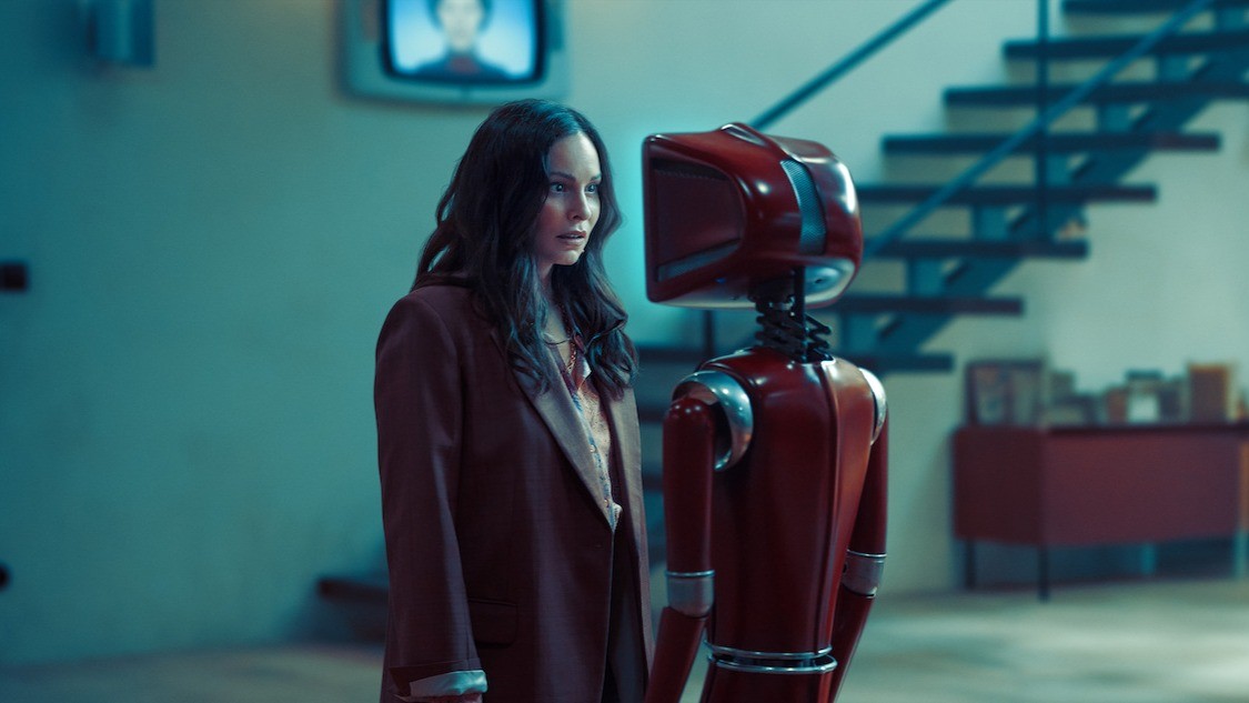 Woman in a burgundy blazer stands beside a red robot in a blue-lit room.