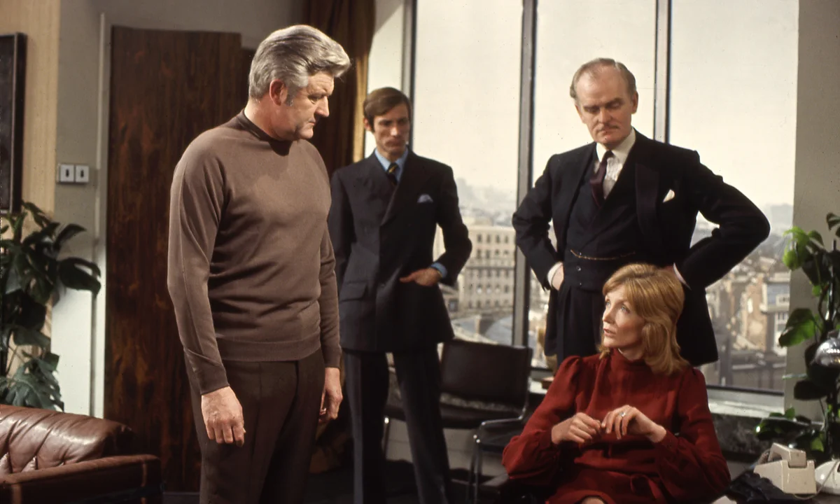 Doomwatch | film.org.pl Man in a brown sweater stands in a modern office, facing a seated woman in red while two suited men observe by a large window.