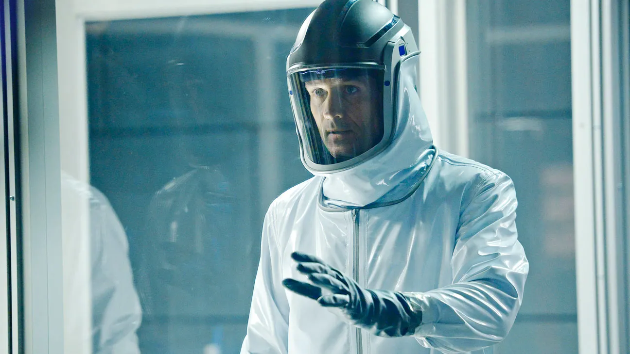 Scientist in a white cleanroom suit and face shield extends a gloved hand in a laboratory setting.