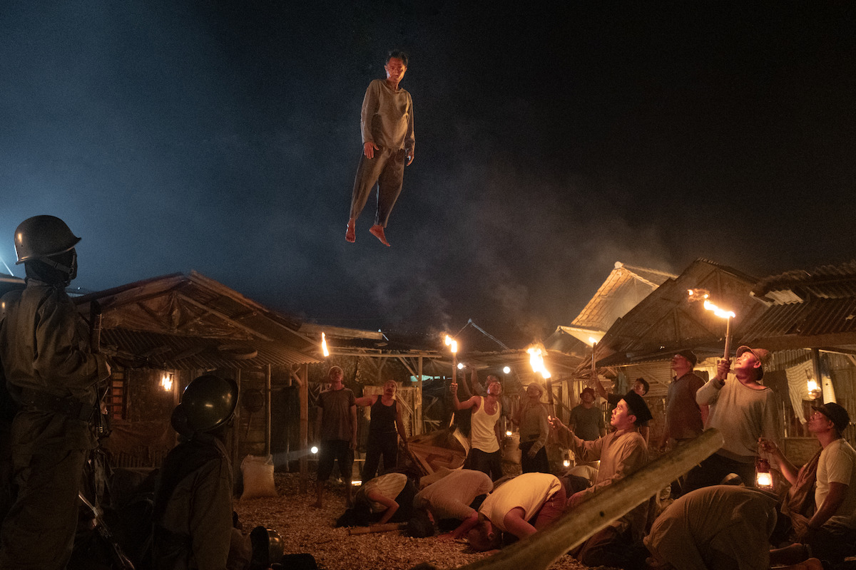 Joko-Anwar-Koszmary-i-sny-na-jawie-2 | film.org.pl Man levitates above a village square at night as a crowd watches with torches surrounding him. Guards stand to the left in protective gear.