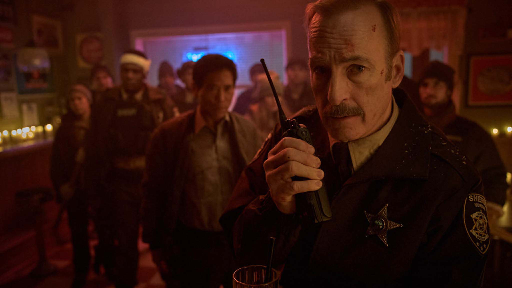 Close-up of a man in a sheriff-like jacket speaking into a radio in a dim, crowded bar scene with blurred patrons in the background.