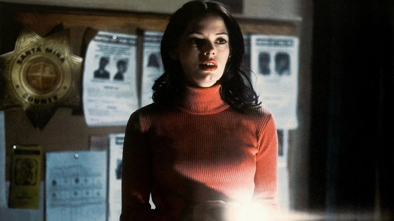 Woman with dark hair in a red turtleneck stands in a dim room, lit from below, with posters and a badge on the wall behind her.
