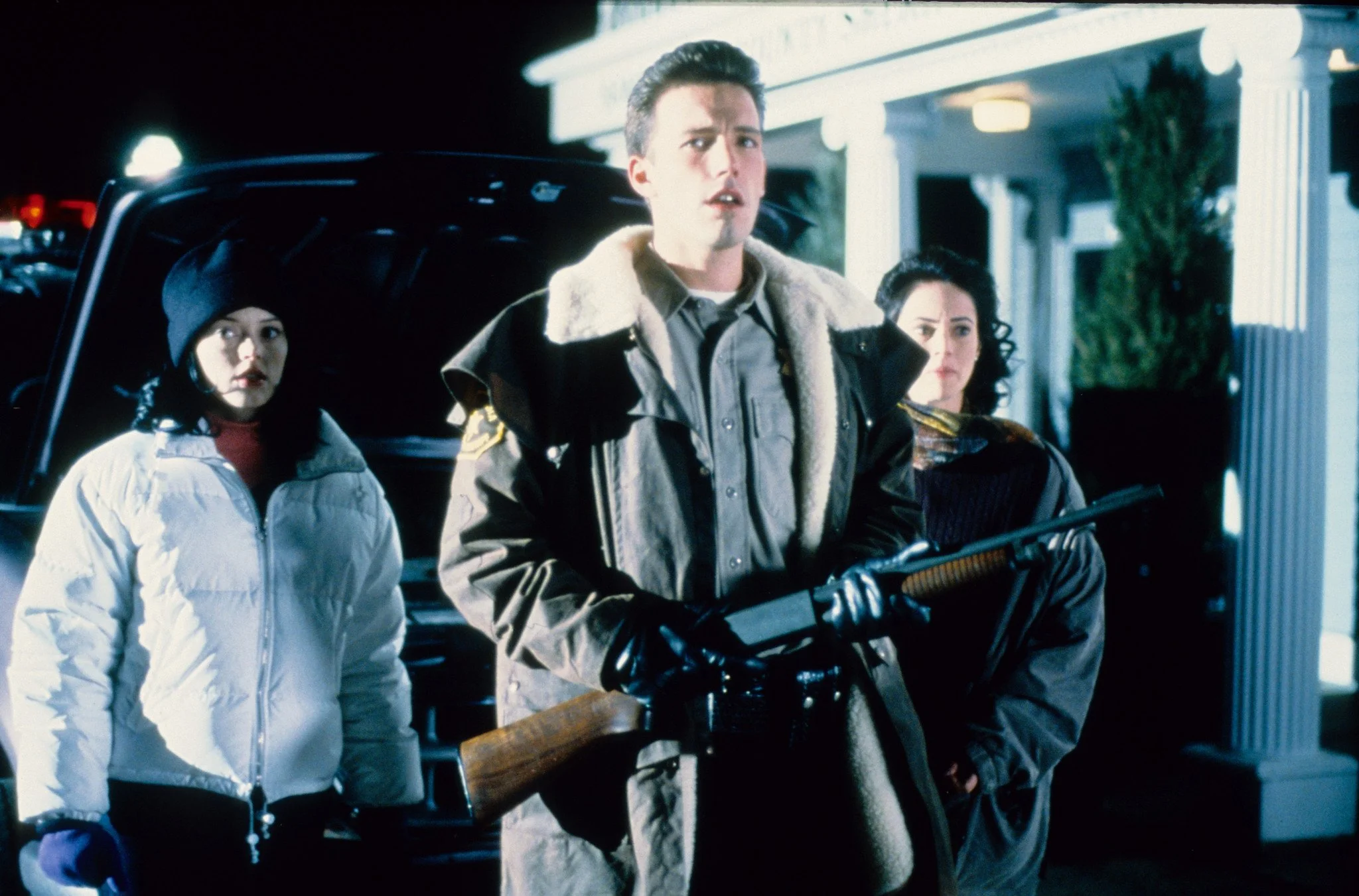 Three people in winter jackets stand outside at night; a man in the center holds a rifle with a wooden stock while two women look on.