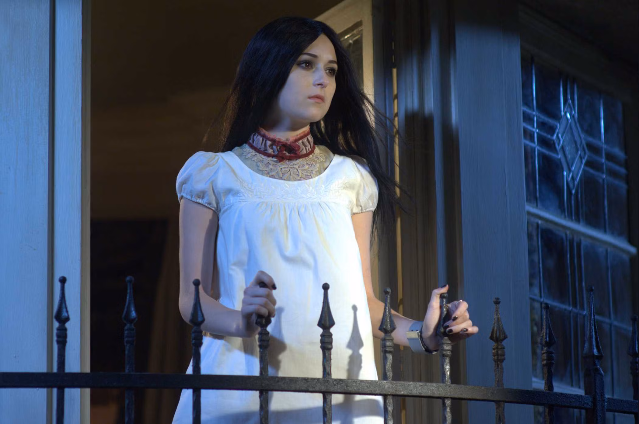 Repo-The-Genetic-Opera-2 | film.org.pl Woman in a white lace-collar dress standing at a balcony, looking pensively to the side at night/beige doorway light.