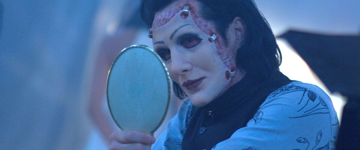 Repo-The-Genetic-Opera-3 | film.org.pl Theatrical performer in a pale mask with red accents and dark hair, peering into a small round mirror under cool blue lighting.