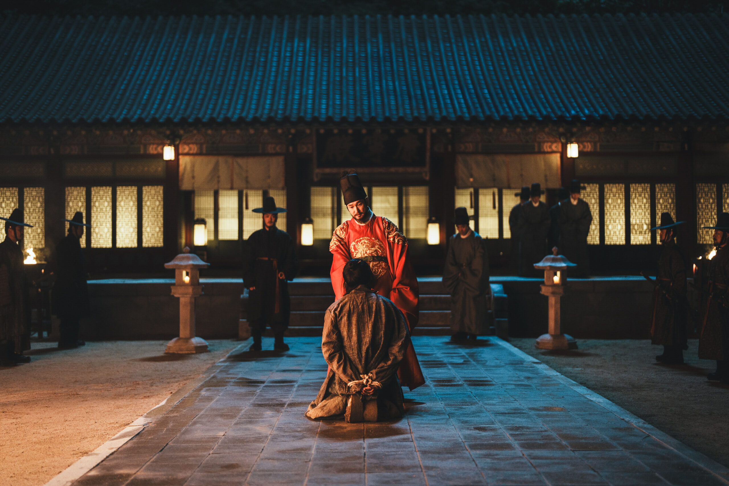 A man in ornate red ceremonial robes kneels before another man in brown robes in a dim, historic courtyard with lanterns and attendants in traditional attire.