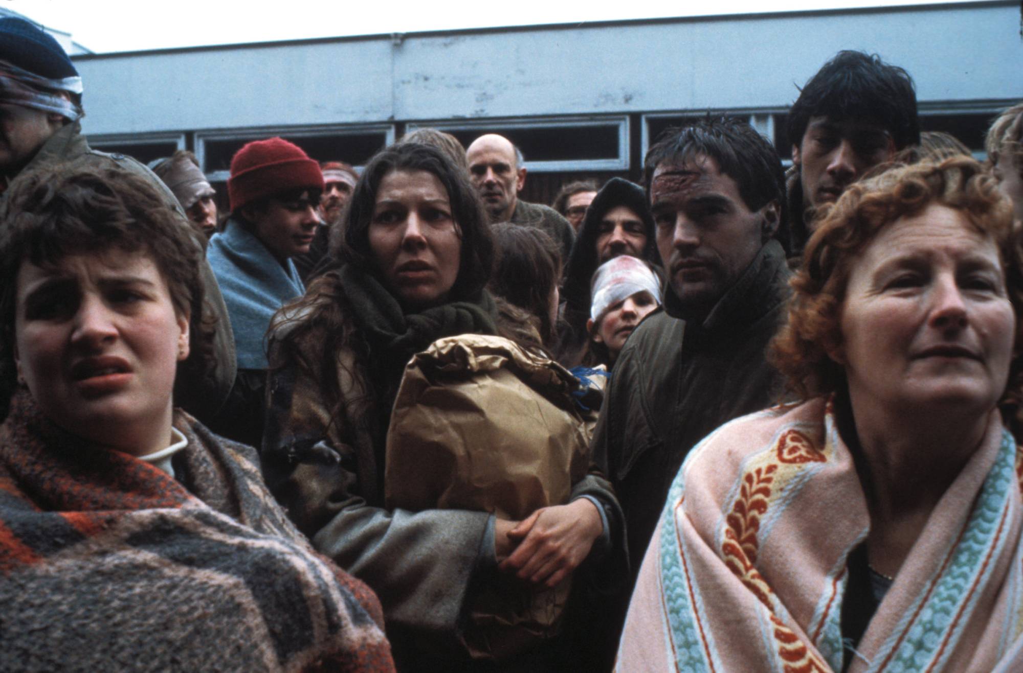 Crowd of adults wearing coats and blankets, looking anxious in an outdoor setting.
