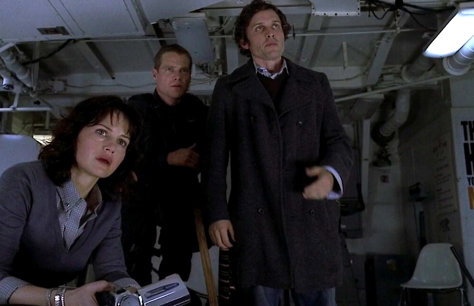 Three people in a dim, industrial control room look intently toward something off-screen; woman seated in the foreground, two men standing behind her.