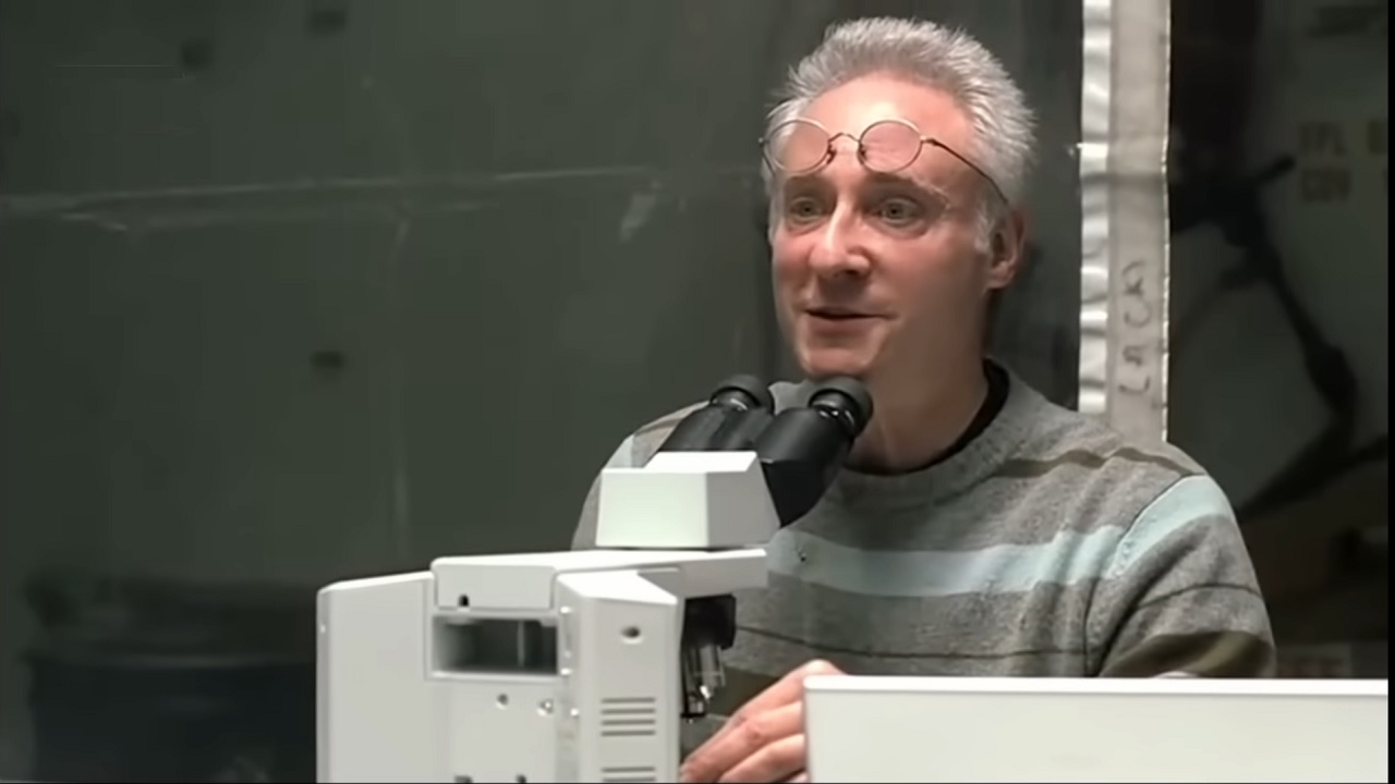 Man with gray hair and round glasses speaks into a microscope setup in a laboratory, wearing a striped sweater.