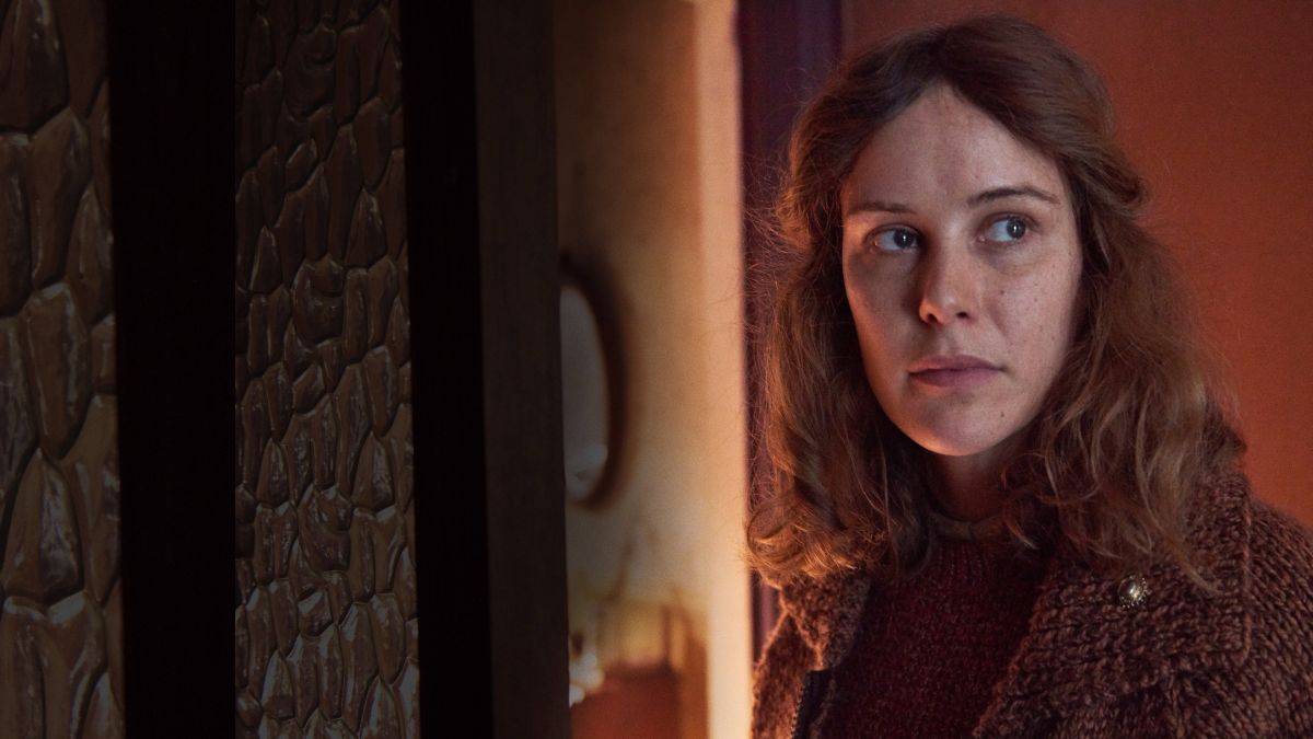 amulet2020 5 | film.org.pl Woman with wavy brown hair in a brown knit sweater stands near a doorway, looking to her left in a warmly lit interior.