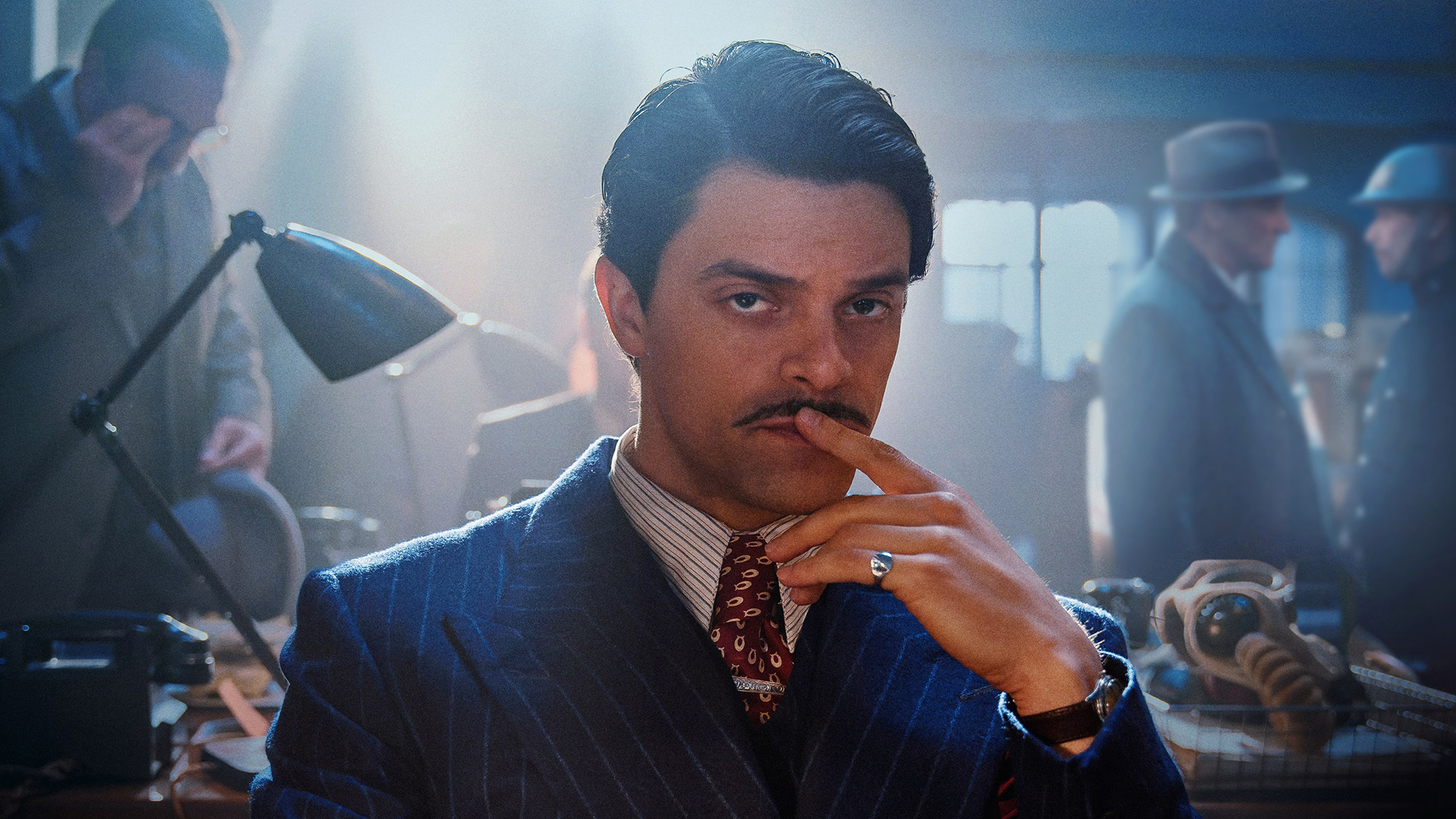 bodies-ciala-2 | film.org.pl Man in a blue pinstripe suit and red tie sits at a desk, finger to lips, in a retro office/film set.