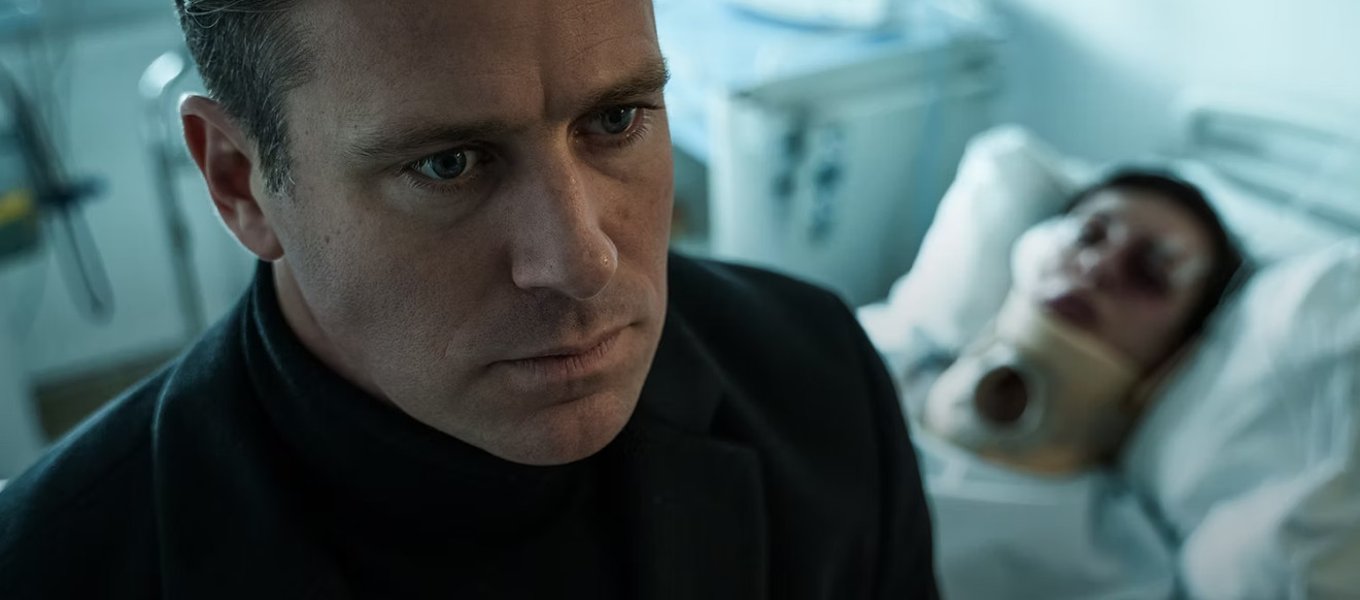 citizen-vigilante-3 | film.org.pl Man in a black turtleneck looks tense as he stands near a hospital bed, with a patient wearing a neck brace in the background.