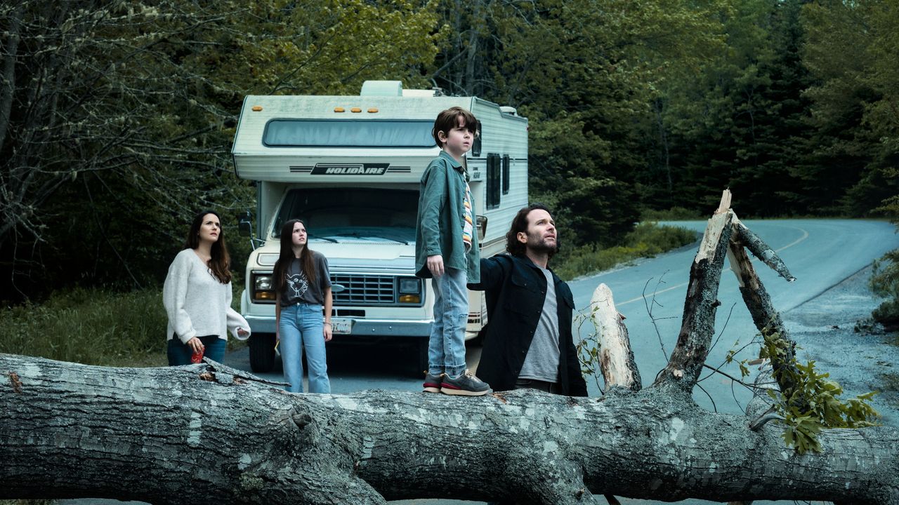 from-stamtad-2 | film.org.pl Family with a camper van on a forest road as a large fallen tree blocks the path; a man holds a boy standing on the log while two women look on.
