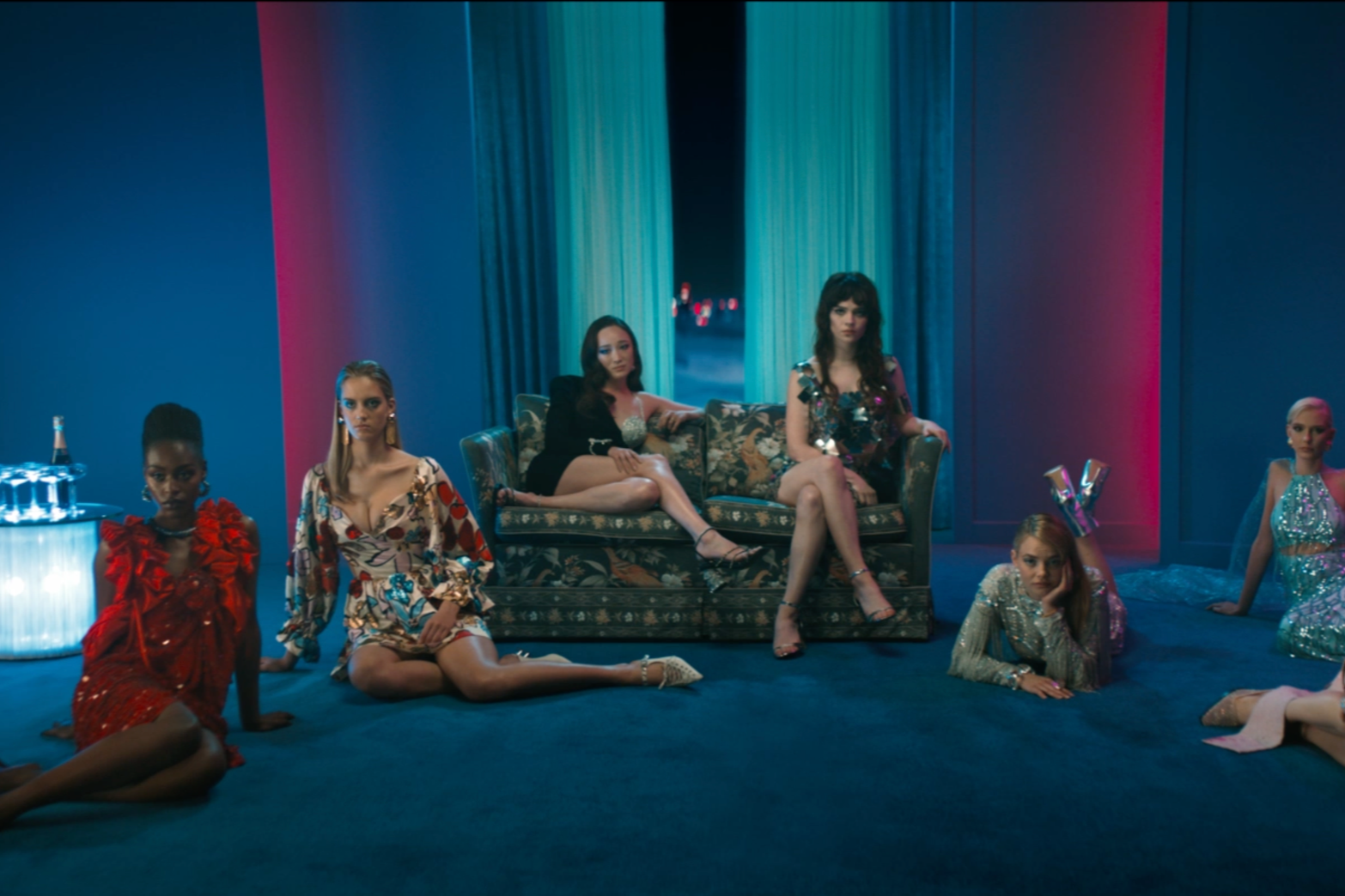 her-private-hell-jej-pieklo | film.org.pl Group of stylish women posing in a neon-lit blue room around a vintage floral sofa, wearing bold party outfits and heels.