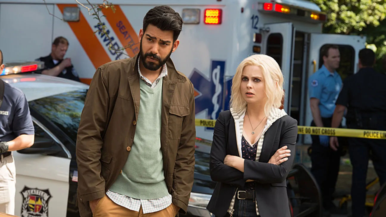 izombie | film.org.pl Two worried civilians stand near an emergency scene: a man in a brown jacket and a green sweater, and a woman with blonde hair in a black blazer, with an ambulance and police tape in the background.