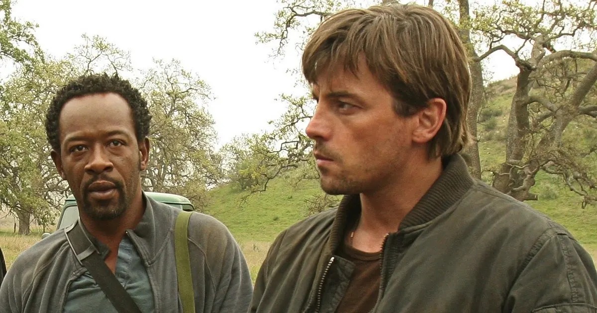 Two men stand outdoors in a grassy, tree-filled landscape, one facing left and the other in profile to the right in jackets.