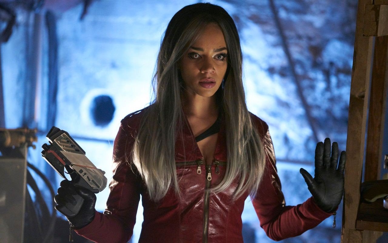 Woman in a red leather jacket holds a futuristic gun, in a blue-lit, industrial setting with a determined look.