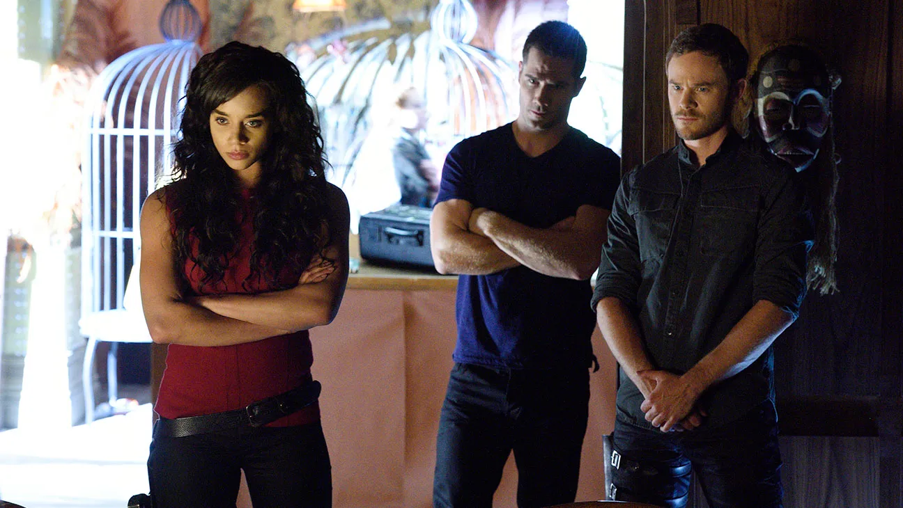 Three adults stand with arms crossed in a dim room: woman in red left, man in navy center, man in gray right; decorative masks hang on the wall behind them.