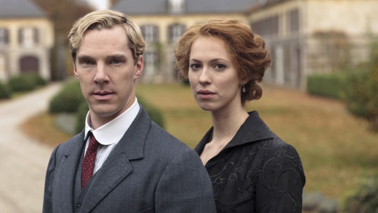 A man in a gray suit and red tie stands in front, with a woman in a black patterned dress behind him, both looking toward the camera in a formal outdoor setting.