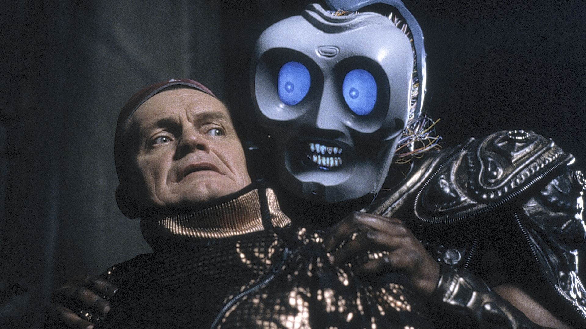lexx-2 | film.org.pl A menacing robot with large blue eyes grabs a worried man in a metallic, sequined outfit.