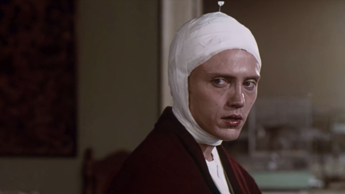 Person with a white head bandage sits in a hospital-like room, looking off to the side.