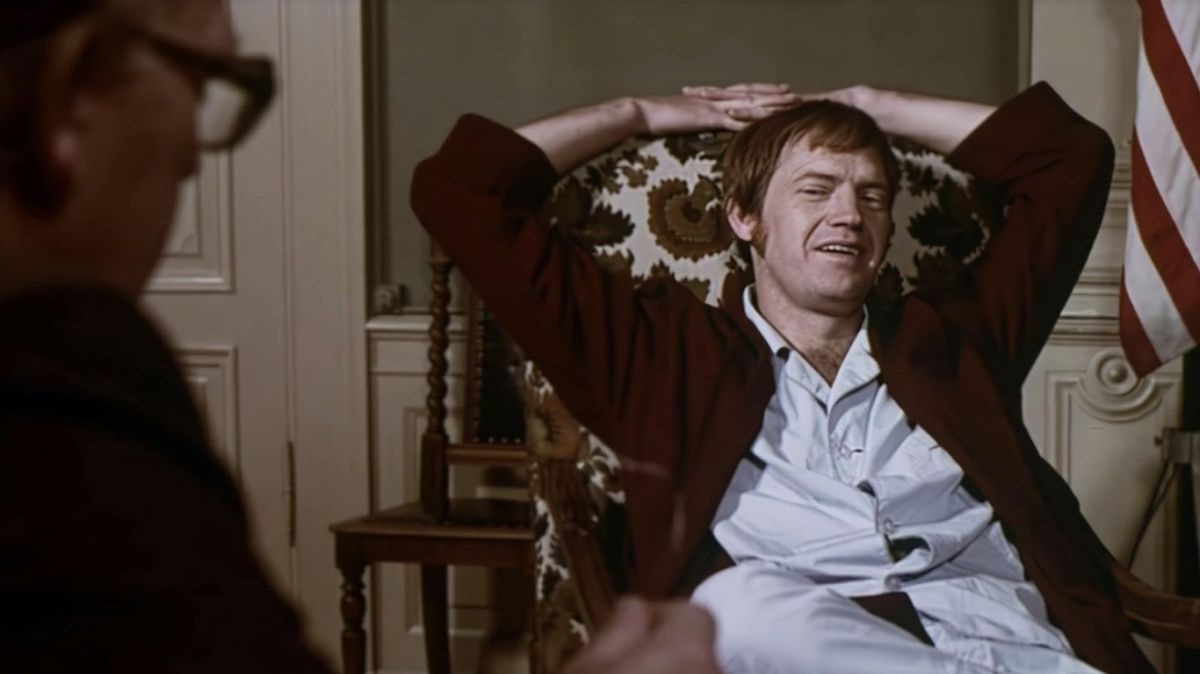 Man in a burgundy robe lounges with arms behind his head in a floral armchair, another person blurred on the left and an American flag in the background.