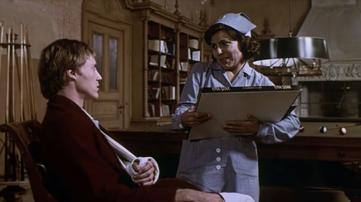 Nurse in a blue uniform and cap shows a clipboard to a seated patient in a vintage hospital room, discussing his care.