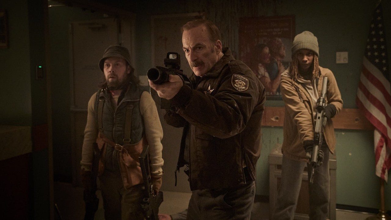 Three armed adults stand in a dim room, aiming their rifles toward the camera, with an American flag in the background.
