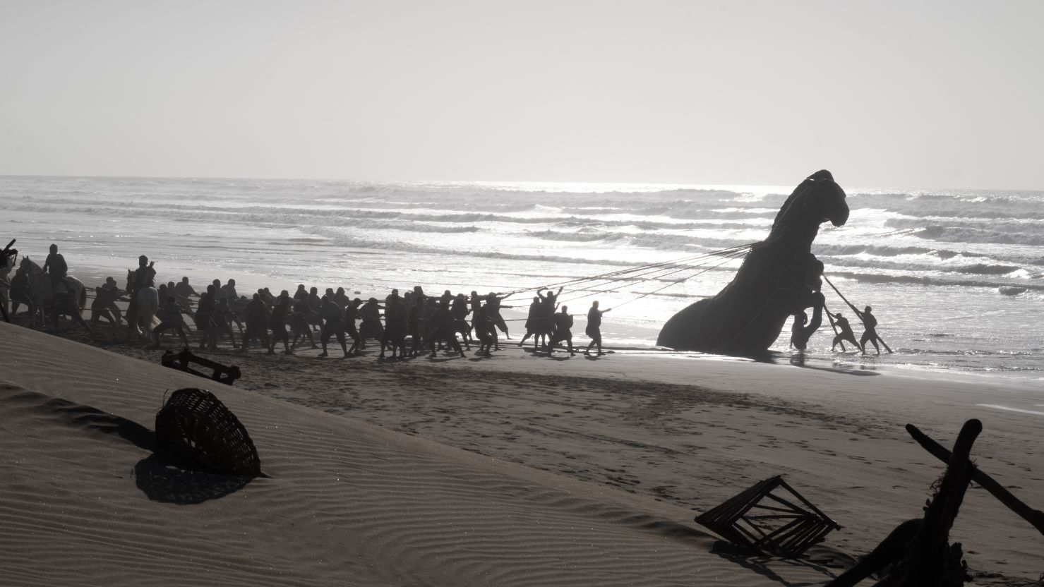 odyssey-2 | film.org.pl Large group of people on a beach pulling a massive whale or sea creature ashore with ropes at sunrise/sunset, breaking waves in the background.